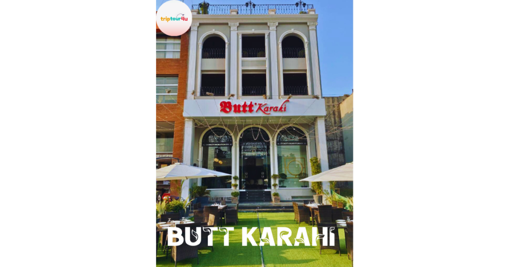 Exterior view of Butt Karahi restaurant with elegant multi-story facade, outdoor seating area with tables and umbrellas, and green lawn entrance under a clear blue sky in Pakistan.