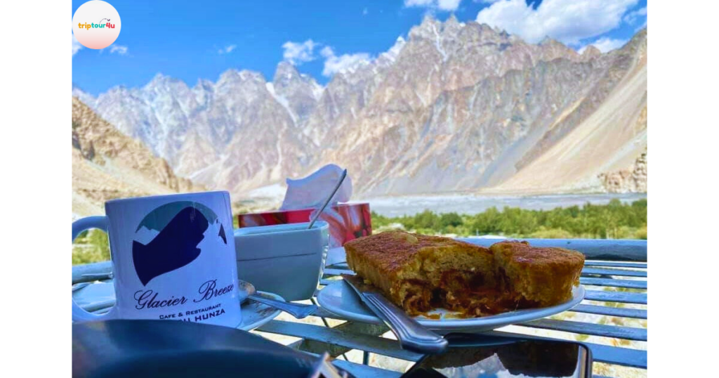 A cup and plate of dessert at Glacier Breeze Restaurant set on an outdoor table overlooking stunning jagged mountains, a bright blue sky, and a lush green valley in Hunza.