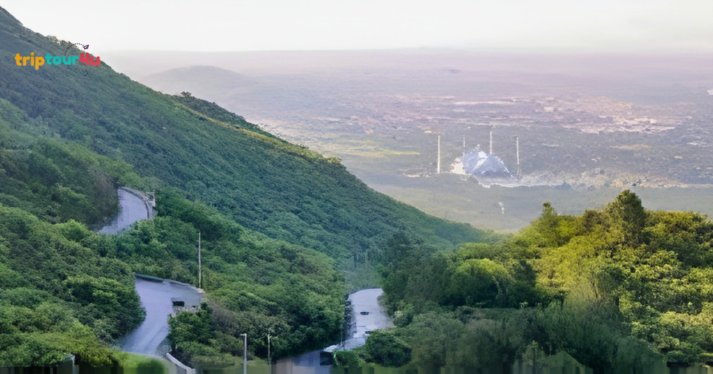 An elevated, high-angle view of a winding asphalt road snaking through a lush, green mountain valley. In the distant, misty background, the white dome and tall minarets of the Faisal Mosque in Islamabad, Pakistan, are visible under a soft, hazy sky. The foreground and midground are dominated by dense, vibrant green foliage and the steep slopes of the Margalla Hills.