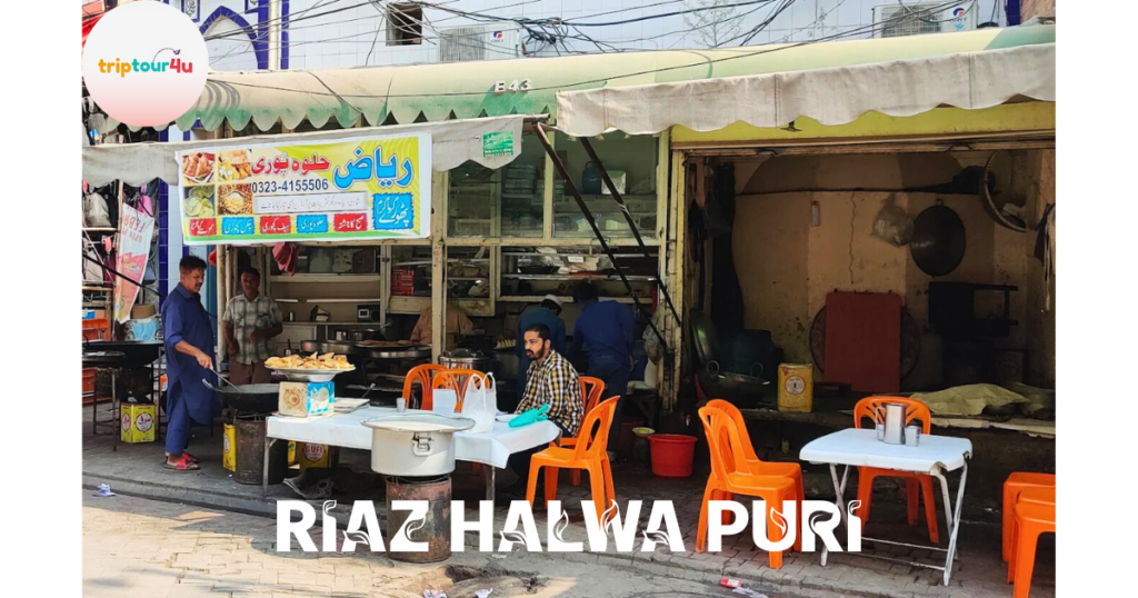 Street view of Riaz Halwa Puri with a traditional roadside setup, cooks preparing halwa puri, orange plastic chairs and tables for customers, and a colorful Urdu signboard in a busy Pakistani market area.