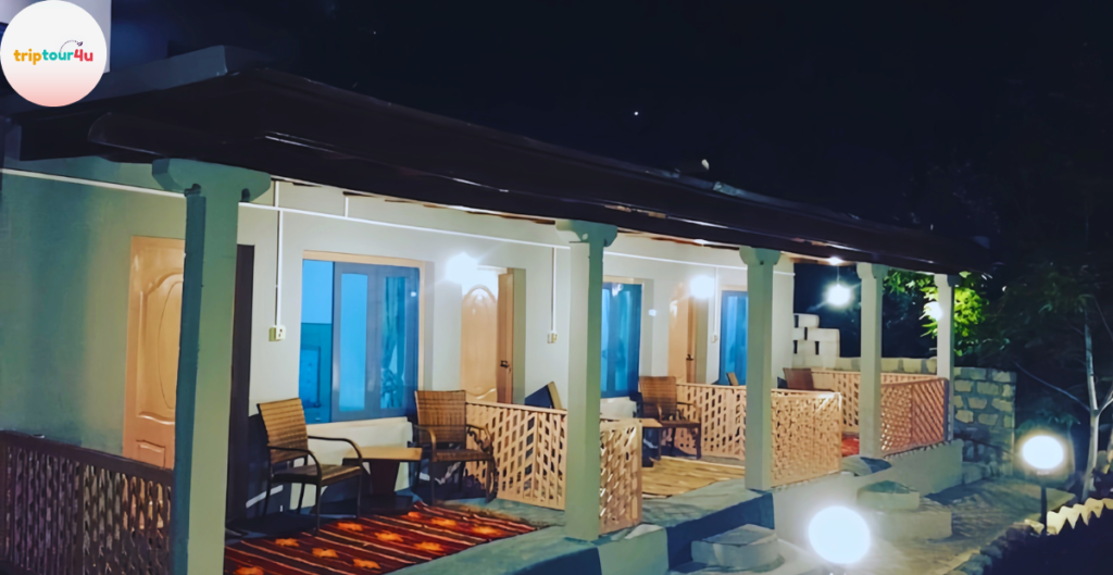 Night view of Hunza Darbar Hotel featuring a row of warmly lit rooms with private terraces, wooden chairs, and decorative railings surrounded by garden lights.