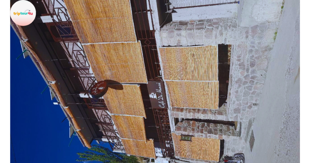 Exterior view of “Cafe De Hunza,” featuring stone walls, wooden shades covering the front, and a signboard hanging above the entrance under a bright blue sky.