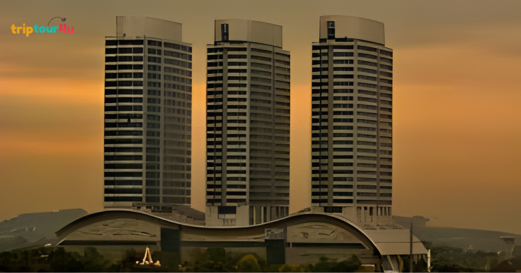 A wide shot of three identical, modern high-rise towers standing side-by-side against a warm, orange-hued sunset sky. The buildings feature a repetitive pattern of rectangular windows and are situated atop a base with a distinctive, wave-like curved roofline