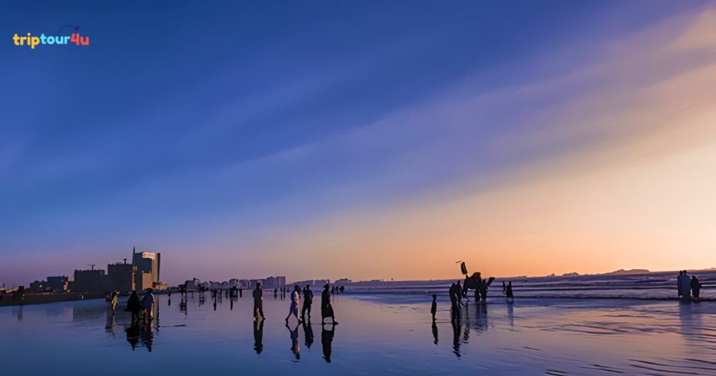 People walking along Clifton Beach, Karachi at sunset, with reflections on wet sand, calm sea waves, and a colorful evening sky.