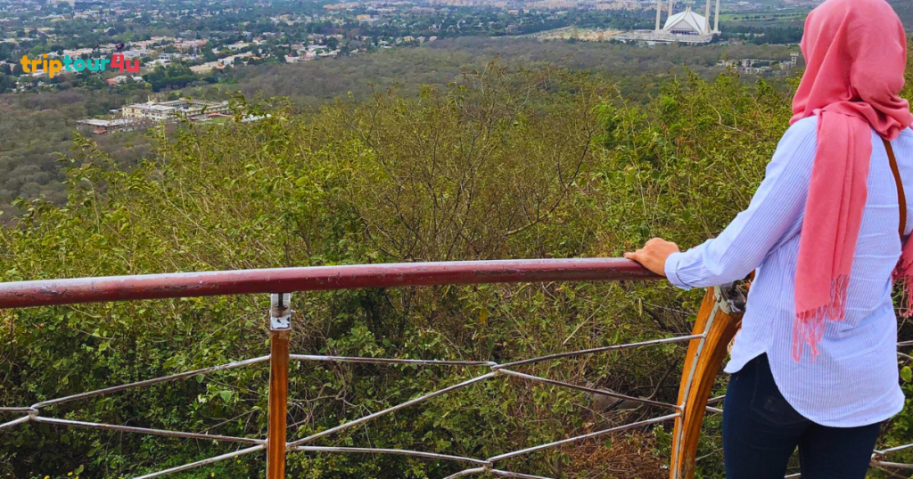 A person wearing a pink headscarf and a striped blue shirt stands at a wooden railing, looking out over a vast green valley. In the distance, the white structure of the Faisal Mosque is visible against the sprawling urban landscape of Islamabad.