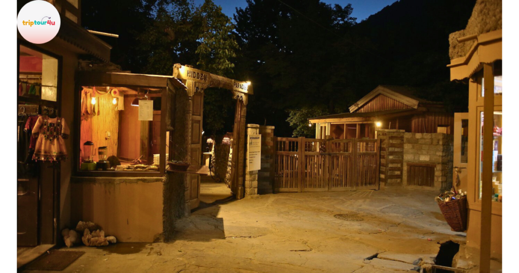 Nighttime view of Hidden Paradise Café, featuring a warmly lit entrance with a wooden archway, surrounding stone and wooden structures, small shops, and a cozy outdoor pathway leading to the café.