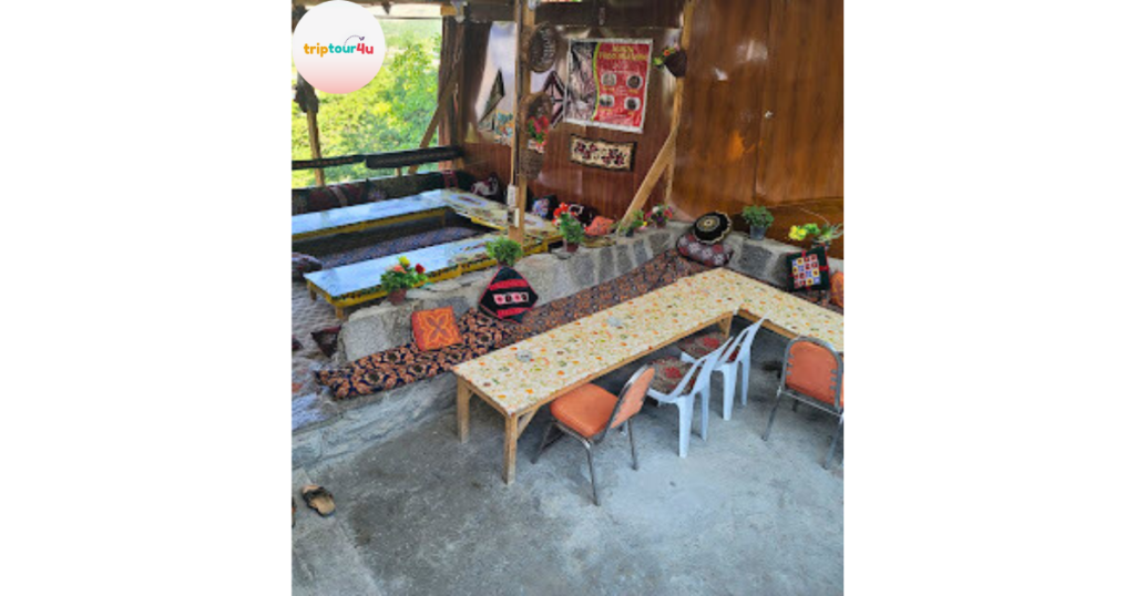 A cozy traditional seating area at Hunza Food Pavilion, featuring long wooden tables, colorful cushions, woven décor, wall hangings, and potted plants arranged around raised stone platforms in an open, rustic setting.
