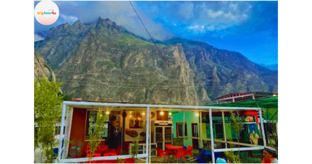 Outdoor view of Hunza Grill Trout restaurant with bright seating under a white-framed structure, set against towering rocky mountains and a dramatic blue sky.