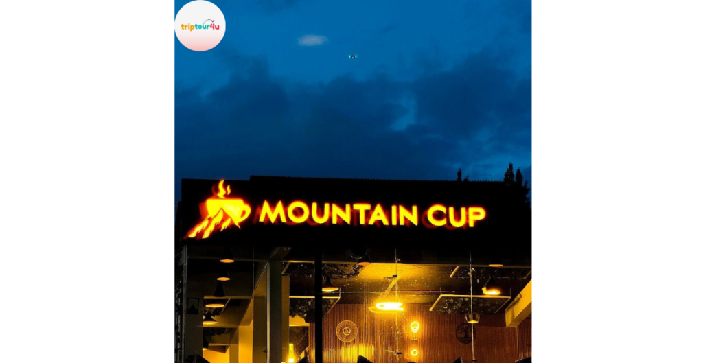 Illuminated “Mountain Cup” café sign glowing in bright yellow-orange against a dark blue evening sky, with warm interior lights visible through the glass below.