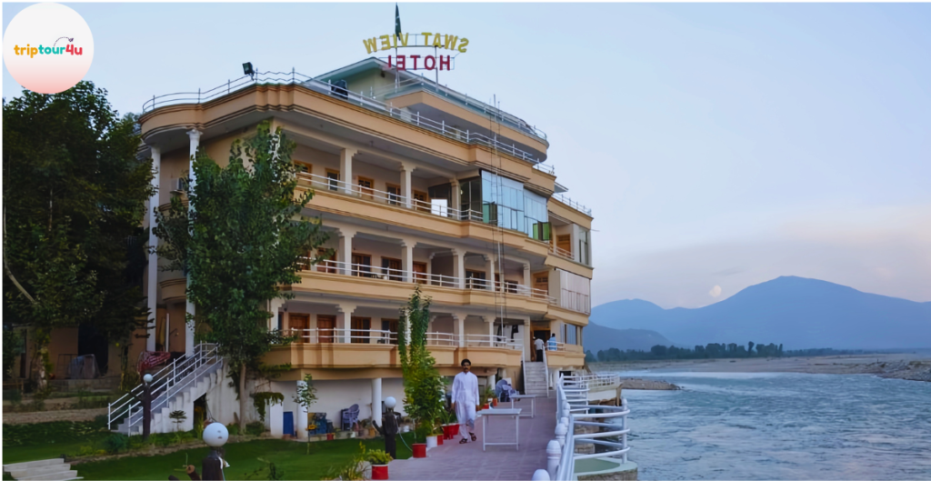 Riverside view of Swat View Hotel, a multi-story building with balconies, cream exterior, surrounding trees, a walkway along the river, and distant mountains under a soft evening sky.