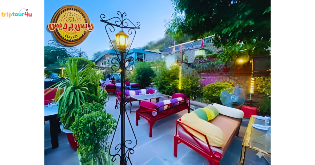 Outdoor restaurant seating area with colorful cushioned sofas, garden plants, and decorative lantern lighting at dusk.