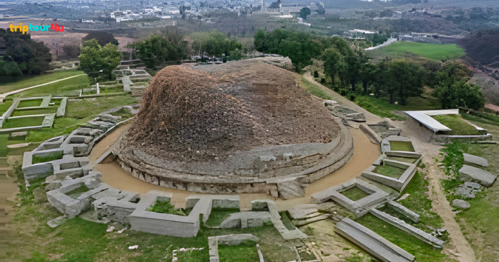 The ancient Dharmarajika Stupa in Taxila, showing a large circular stone mound surrounded by ruined platforms, pathways, and green landscape in the background.
