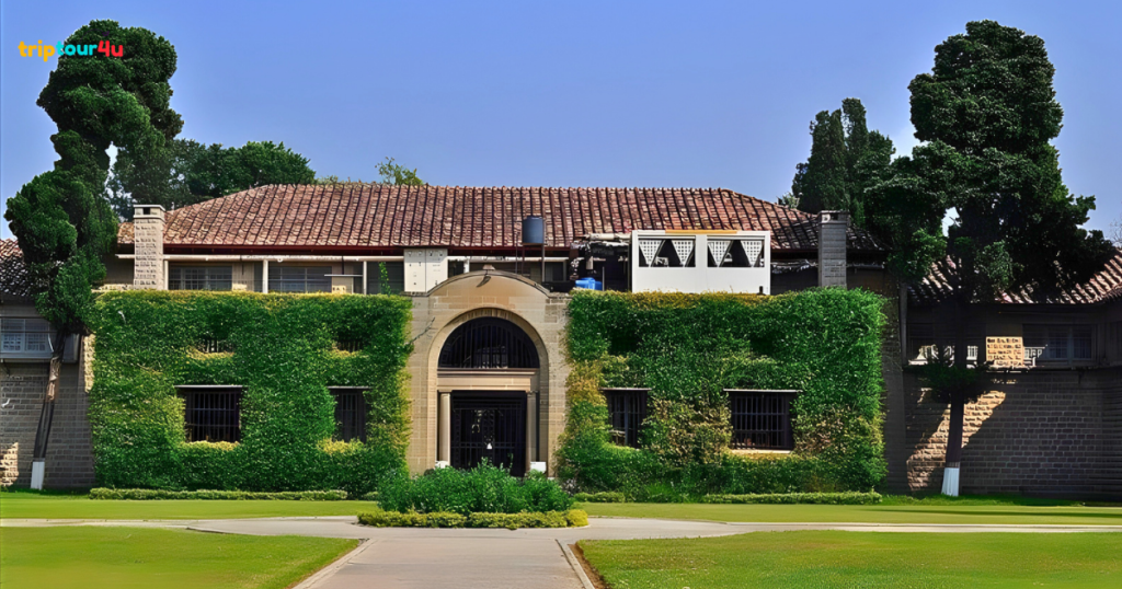 Front view of a historic stone building with an arched entrance, red tiled roof, ivy-covered walls, and manicured green lawn, framed by tall trees under a clear blue sky.