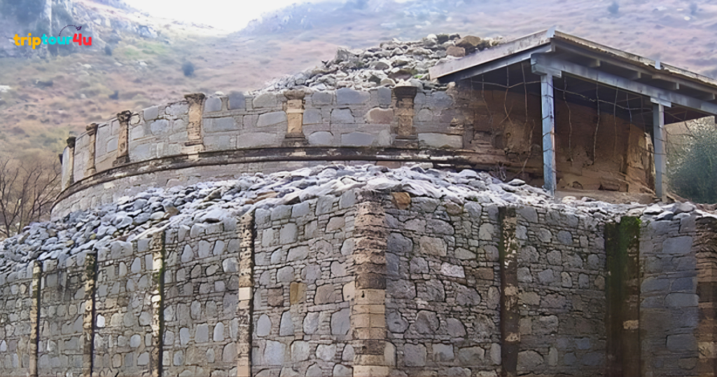 Circular stone stupa ruin with layered masonry walls and a partially collapsed upper structure, set against a mountainous landscape, showing ancient Buddhist-era architecture in weathered condition.