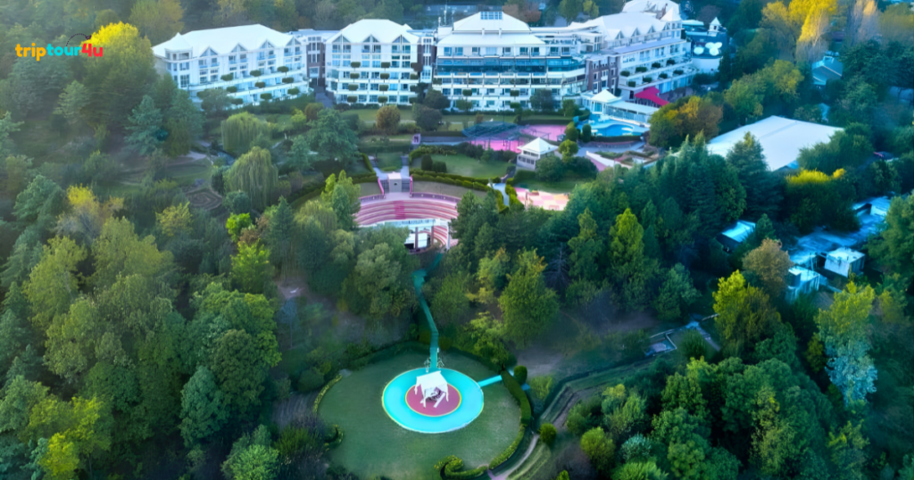 “Aerial view of a large hilltop resort complex surrounded by dense green forest, with white buildings, landscaped gardens, an outdoor amphitheater, and a circular helipad connected by winding paths.”