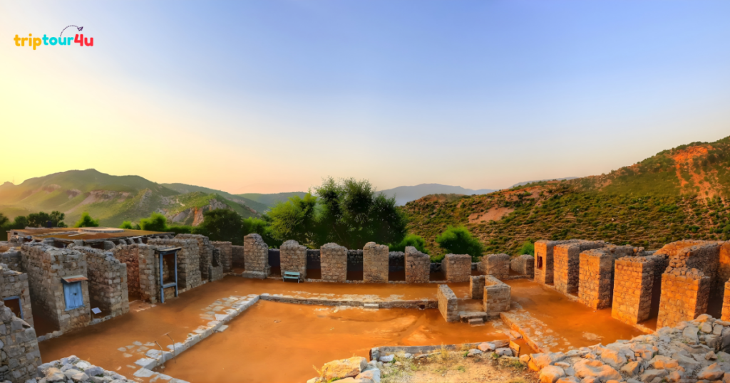 Ancient stone ruins at the Julian Monastery in Taxila, featuring preserved walls and foundations set against green hills and a clear sky at sunset.
