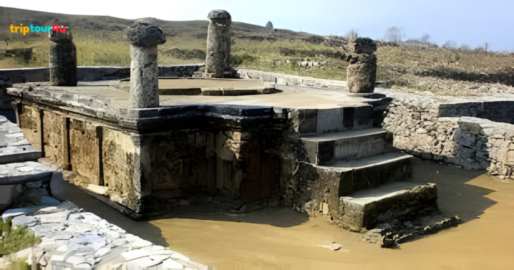 Ancient stone ruins at Sirkap, Taxila, showing a raised platform with weathered pillars, steps, and surrounding water against a hilly landscape.